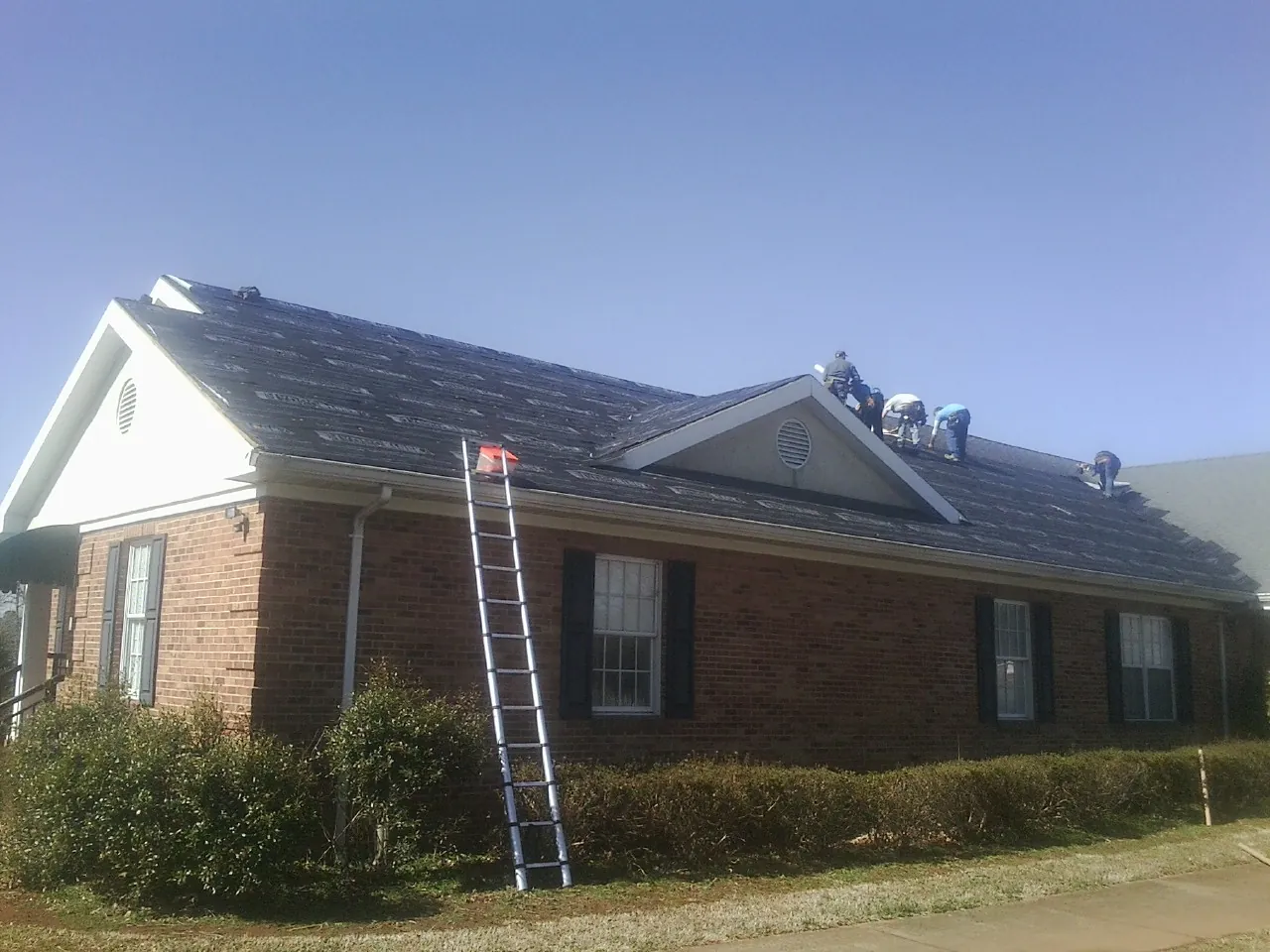 Crew tearing shingles off a brick ranch roof with a ladder set — Metal Roof Repair work by our crew in Lester Prairie