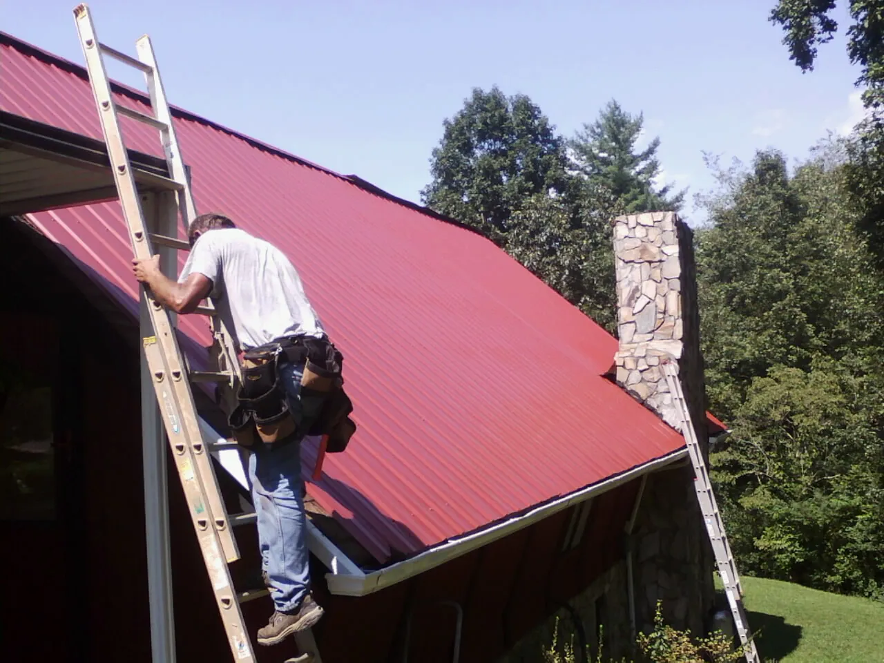 Roofer on a ladder beside a red metal roof and stone chimney — Soffit & Fascia Repair by Roofing Brothers in Lester Prairie